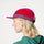 A side-profile view of a woman wearing a 5-panel cap with colour-blocked pink, red, and purple panels, a green hoodie, and hoop earrings against a plain white background.