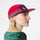A woman in profile wearing a two-tone pink and purple 5-panel cap with a visible Cotopaxi logo on the front panel, set against a plain light grey background.