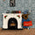 A miniature felt dog character wearing a blue robe and hat, holding a red sign that says 'stories', posed next to a white fireplace decoration with holly and books on a miniature shelf, set against patterned wallpaper.
