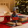 A festive felt ornament shaped like a dog wearing a blue nightshirt and hat, holding a red sign that says 'Stories', styled on a bed with books, a red stocking, and a blurred Christmas tree in the background.