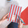 A top-down view of a person holding a striped 2027 diary with red and light blue vertical stripes. The person is holding the diary with both hands, with teal painted fingernails, and holding a matching teal pen.
