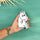 A hand holds a white, Moomin-style character-shaped item with black line art against a teal background with a decorative sun in the corner.