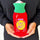 A person in a black sweatshirt holds a red novelty hot water bottle cover designed to look like a hot sauce bottle, with a green ribbed neck and a yellow circle reading 'Shit Hot' in purple.