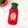 A red knitted hot water bottle with a green neck and a yellow circular graphic that reads 'Shit Hot' in purple, placed on a white faux fur surface with a pink planter partially visible in the corner.