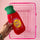 A hand holds a red novelty hot water bottle with a green ribbed neck and a yellow circular label reading 'SHIT HOT' against a pink background, with a pink wire shopping basket behind it.