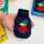 A hand holds a navy blue reusable mini hand warmer featuring an embroidered Woodstock wearing a red coat and green hat. The background shows a white surface with partial Peanuts book covers and the product box.